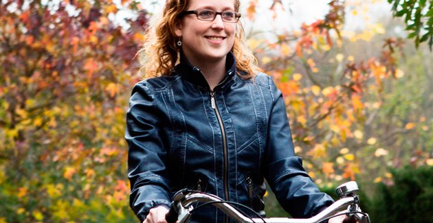 Glimlachende vrouw fietst tussen bomen met bladeren in herfstkleuren.